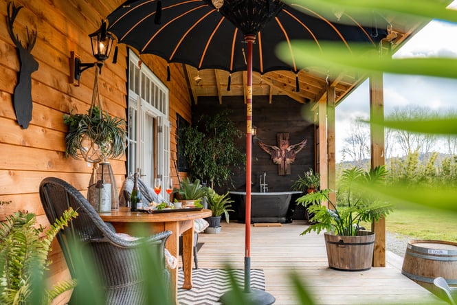 Porch at country house accommodation with an outdoor bath, plants and dining table with cheeseboard and champagne for two