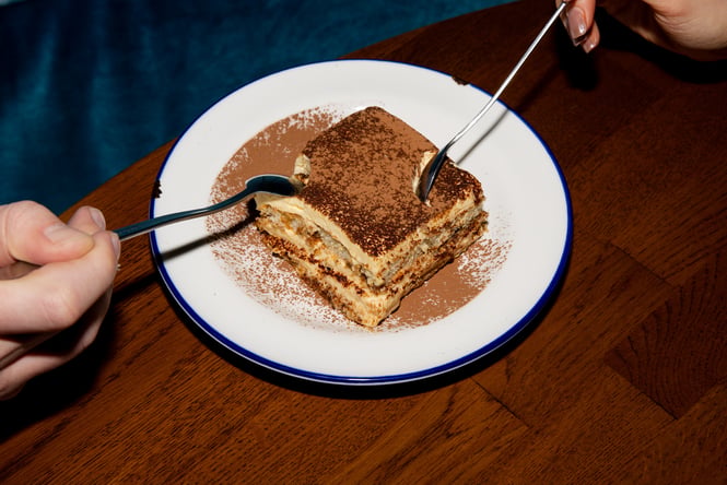 Close up of two hands digging into a plate of tiramisu with a spoon