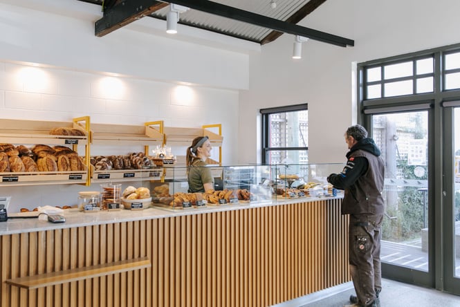 Woman serving man at Bellbird Bakery, surrounded by a fully stocked shop of bread and classic french pastries