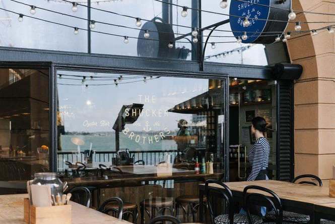 A staff member dressed in blue and white walking in front of the Shucker Brothers restaurant on a sunny day.