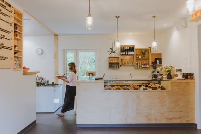 Woman walking with a tray of goods behind the counter and cabinet area at Augustine's of Portobello in Dunedin