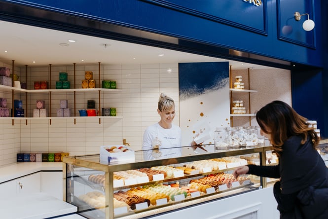 Customer looking at cabinet of macarons with a smiling lady serving her behind the counter
