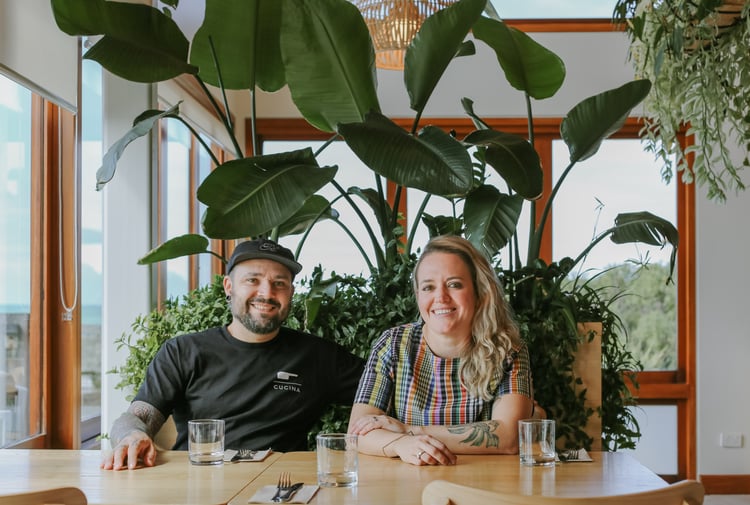 Yanina and Pablo sitting at a table under a plant, smiling to camera.
