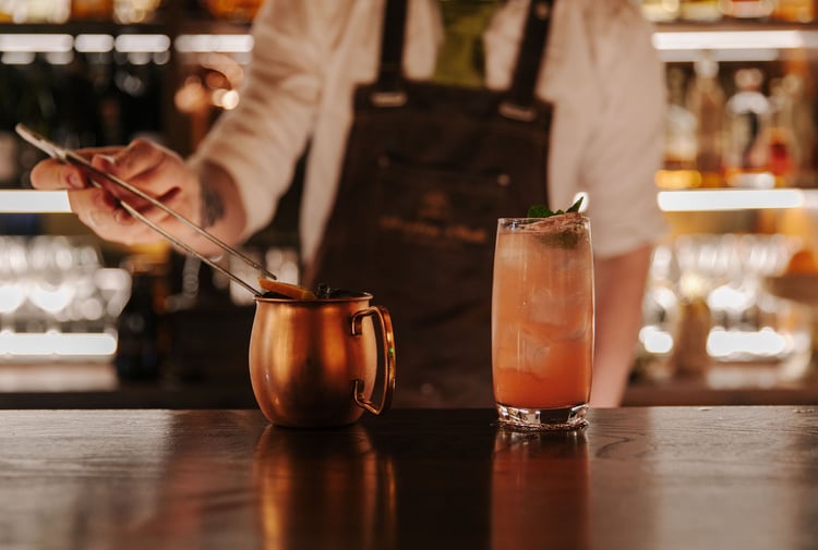Bartender adding a citrus garnish to cocktail in a copper mug next to a pink cocktail with a mint garnish