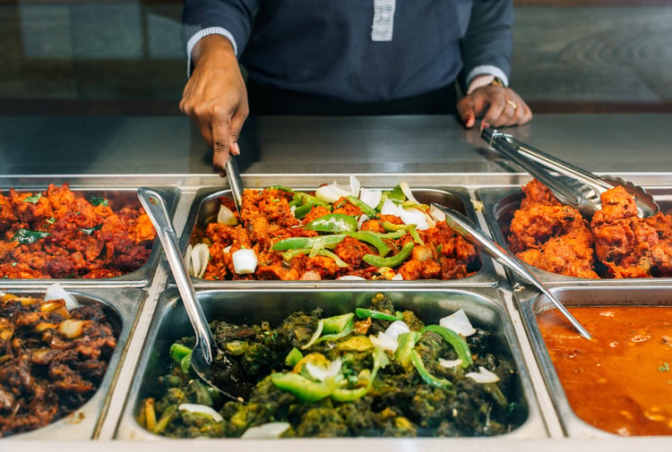 A staff member scooping up curry from a buffet behind glass.