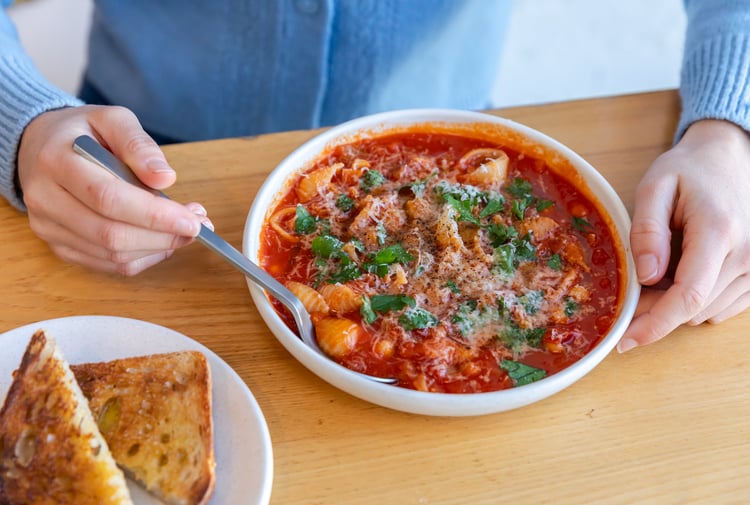 Close up of someone dipping spoon into minestrone soup from Estelle next to plate of toast