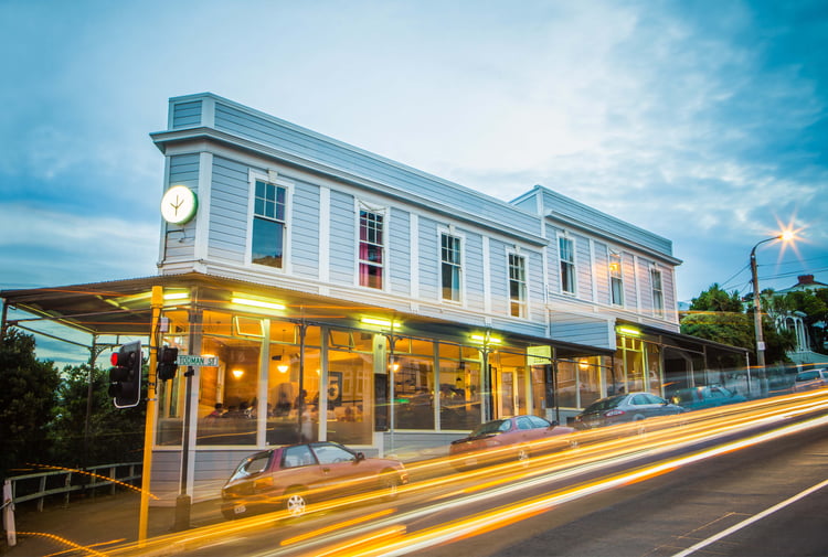 A photo of a blue building with cars whizzing by out front at dusk.