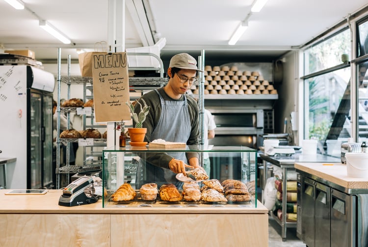 Worker behind cabinet serving almond croissant onto plate using tongs.