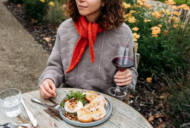 Woman sitting at an outdoor table with a glass of wine in her hand and dish of food in front of her on the table.