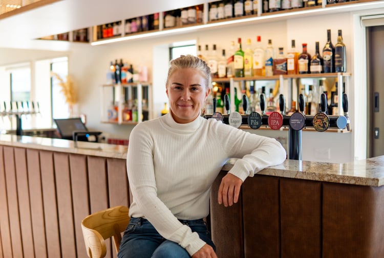 A blonde woman sitting at a bar smiling to camera.