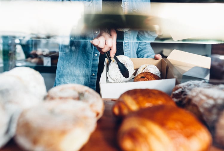 Person in denim jacket using tongs to put donuts and croissants into a take away container.