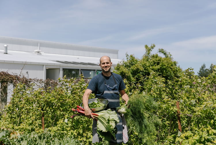 Bevan Smith from Riverstone Kitchen wearing a blue t-shirt and apron smiling to camera in amongst green trees.
