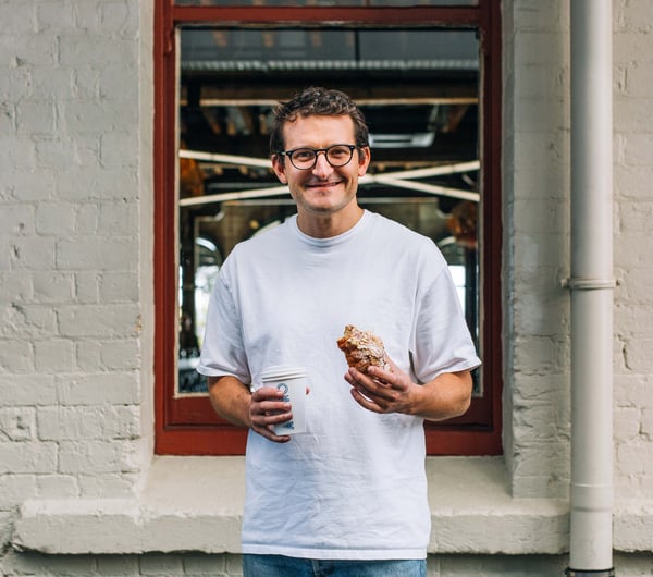Ben Lenart holding a pastry and drink and smiling to camera.