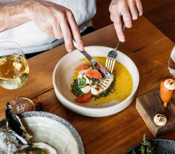 High angle view of a person cutting into a fish and citrus dish, wine and other dishes on the table at arc, Wānaka.