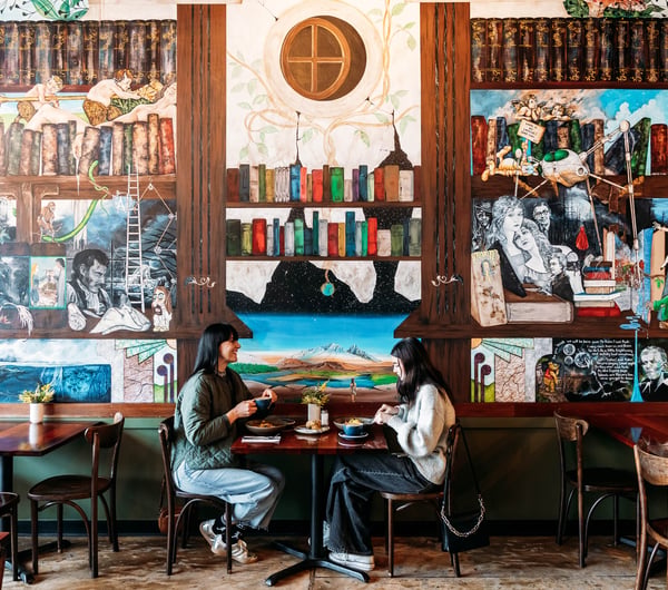 Two diners sitting at a table eating with lots of decorative artwork behind them.