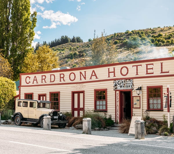 Historic cream and maroon weatherboard frontage of Cardrona Hotel with a vintage cream 1968 Chrysler Model 62 parked in front.