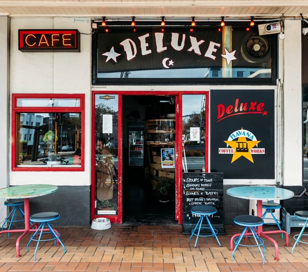 The exterior of Deluxe with its red-framed door and window and round tables and chairs set out on the brick-paved footpath.