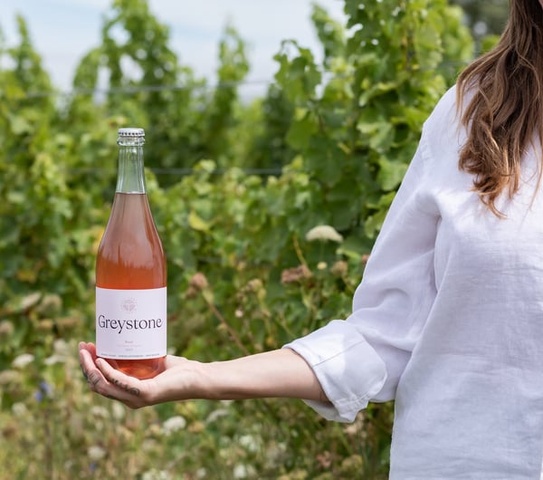 A woman in a white shirt holds a bottle of Greystone Rose Pet Nat against the vineyard's green backdrop at Greystone Wines.