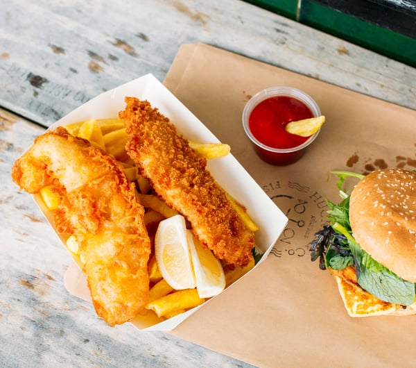 A flatlay of fish and chips and burger with a little pottle of sauce on an outdoor table.
