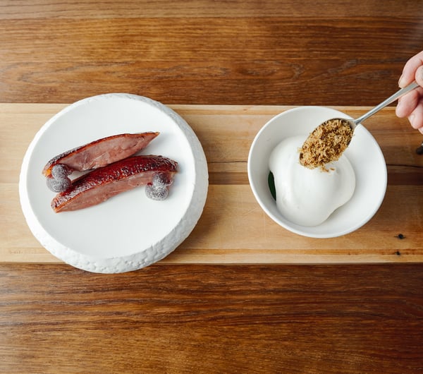 A hand spoons a garnish onto a dish on a wooden board.