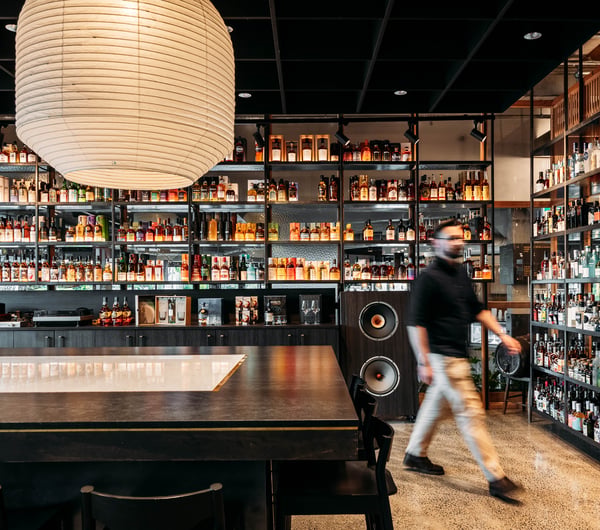 Man walking through the store with bar area in front of him and shelves of alcohol behind him.
