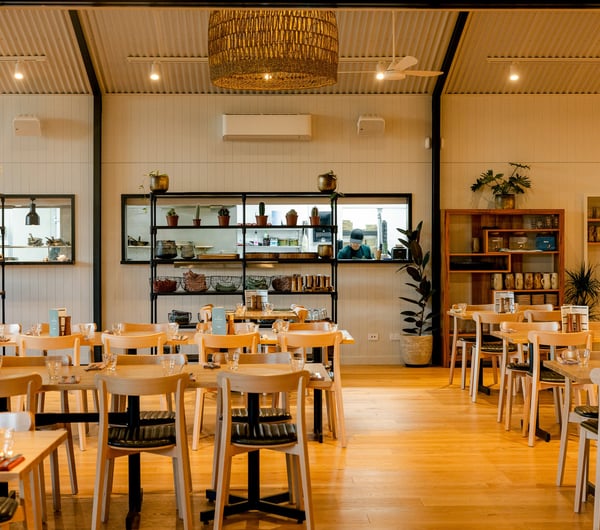 Contemporary and plant-filled interior of Off The Track restaurant with the light wooden dining tables and a chef cooking through the open kitchen window