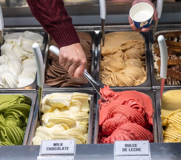 Various different gelato flavours are displayed in a fridge, shop assistant scooping from one flavour into a cup at Paleta Gelateria.