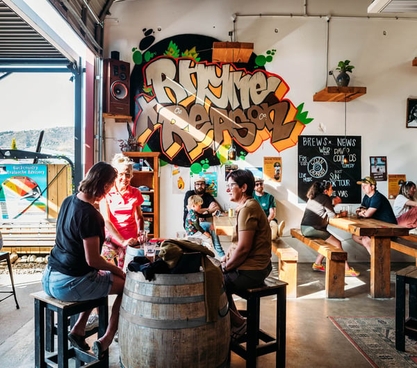 Groups of people sitting inside drinking beers in the sunshine in the high-ceilinged Rhyme and Reason taproom in Wānaka.