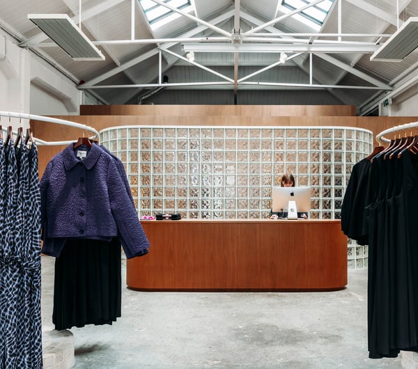 Shop assistant behind the counter with clothing racks in front of her in twenty-seven names.