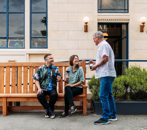 Waiter serving wine to two guests sitting on bench chair outside wine bar