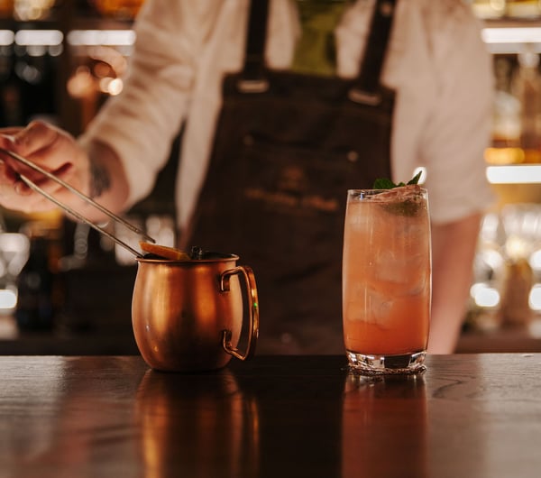 Bartender adding a citrus garnish to cocktail in a copper mug next to a pink cocktail with a mint garnish