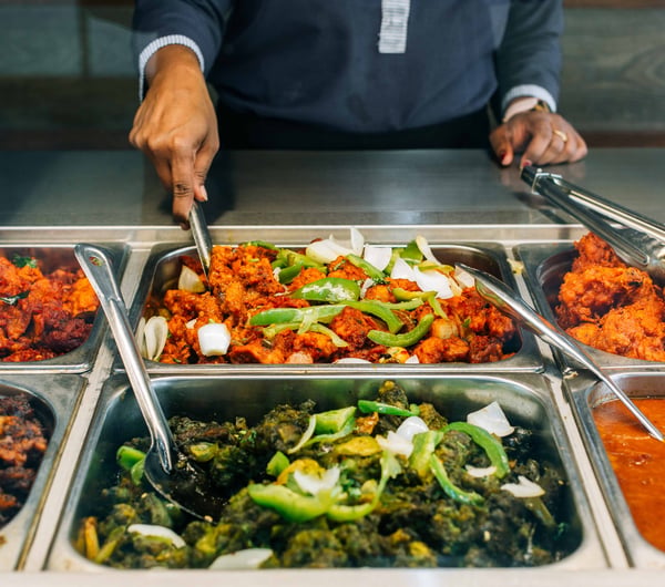 A staff member scooping up curry from a buffet behind glass.