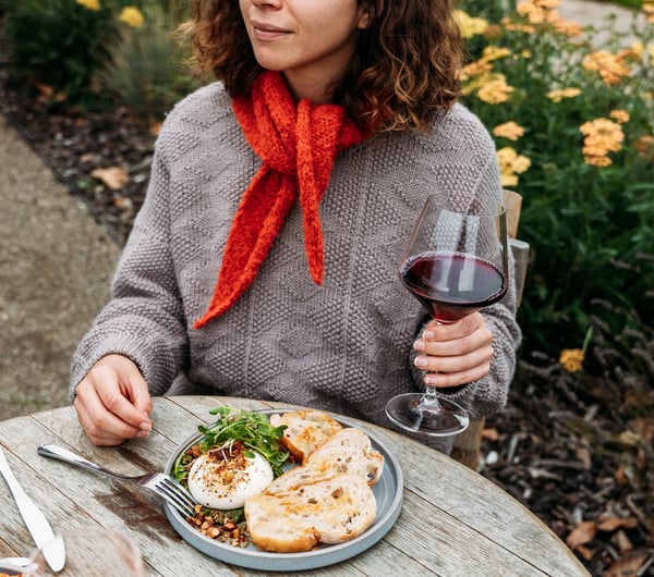 Woman sitting at an outdoor table with a glass of wine in her hand and dish of food in front of her on the table.