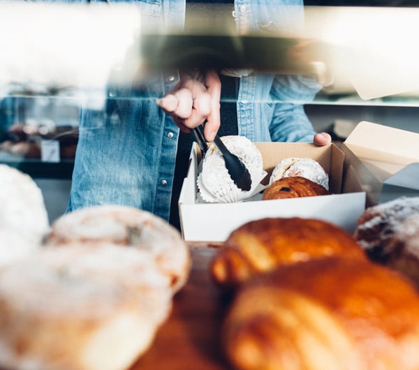 Person in denim jacket using tongs to put donuts and croissants into a take away container.