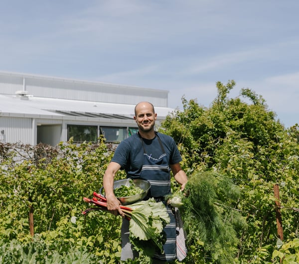 Bevan Smith from Riverstone Kitchen wearing a blue t-shirt and apron smiling to camera in amongst green trees.