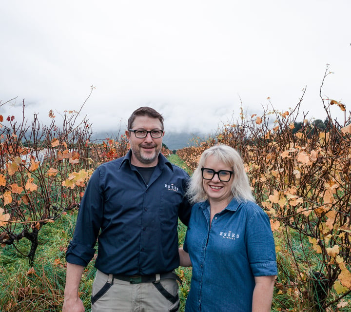 A man and woman smiling to camera in a vineyard on an autumn day.