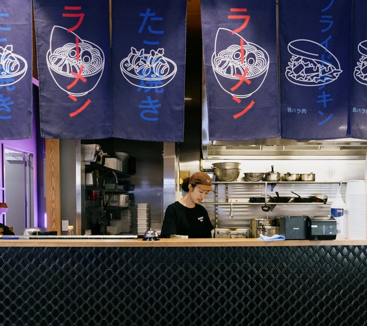 Worker standing behind counter and under Japanese style flags at Ramen Ria in Christchurch
