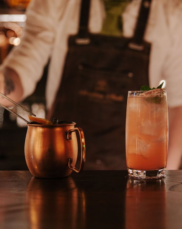 Bartender adding a citrus garnish to cocktail in a copper mug next to a pink cocktail with a mint garnish