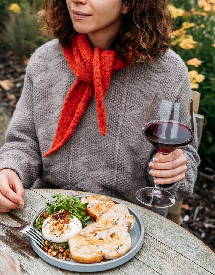 Woman sitting at an outdoor table with a glass of wine in her hand and dish of food in front of her on the table.