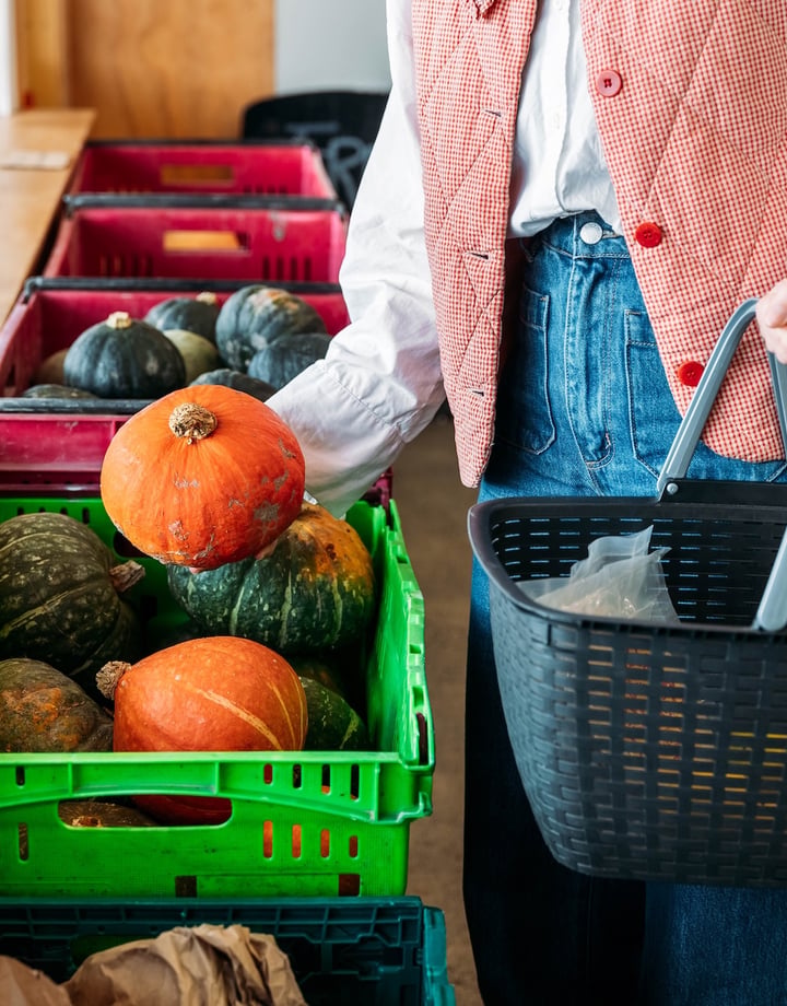 A person holding a shopping basket picks an orange pumpkin out of a plastic crate.