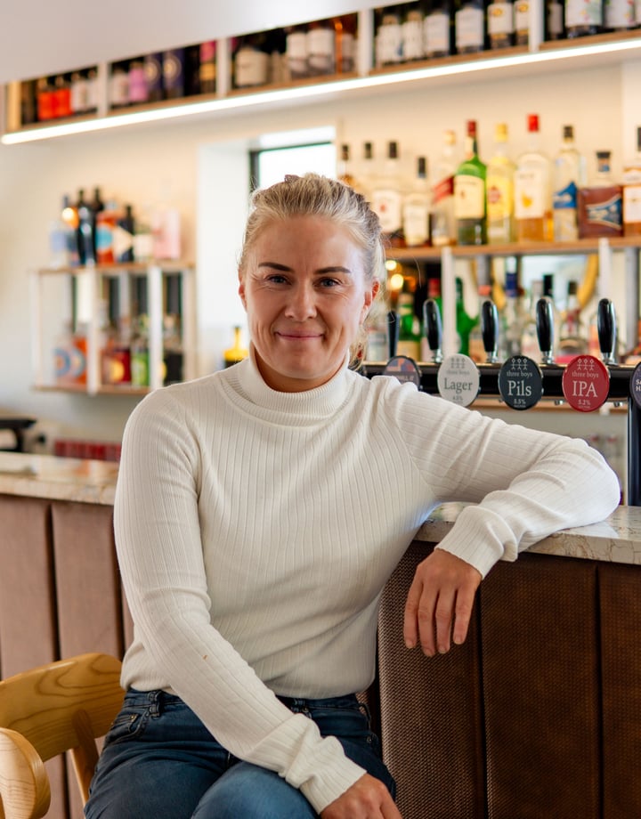 A blonde woman sitting at a bar smiling to camera.
