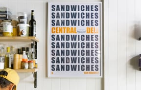 A large Central Deli Sandwiches poster framed on a wall.