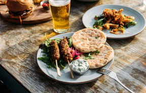 A plate of lamb koftas, flatbreads and tzatziki on a table at hello RANGER with a pint of beer and other plates in the background.
