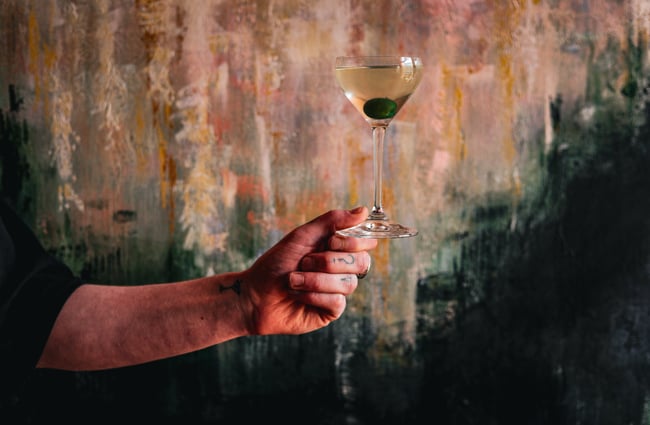 An outstretched arm holds the base of a cocktail; a martini with an olive in it, at gin gin.