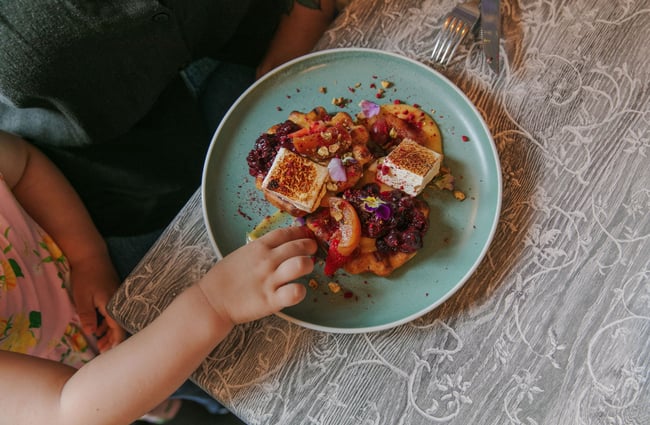 Close up of a toddler's hand reaching to take fruit off the plate of sweet waffles.