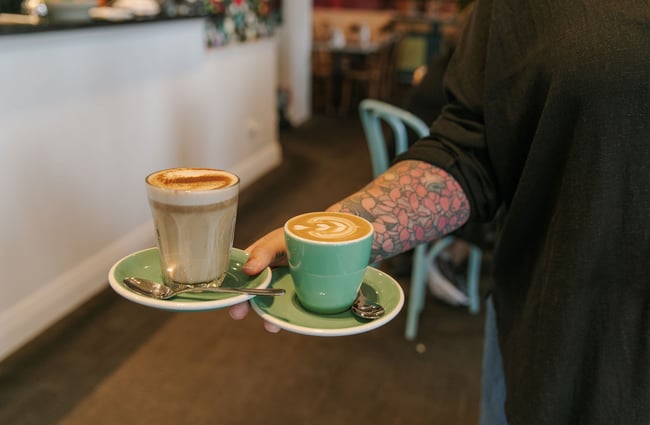 Close up of a waiter holding two coffees balanced on green saucers in their right hand.
