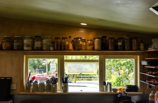 Jars of spices on a high shelf and utensils in jars on a windowsill inside the kitchen prep area at Okere Falls Store.