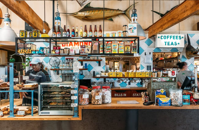 The counter at Seashore Cabaret with a full pie warmer, baked goods, jars of cookies, hanging shelves with goods for sale, a blue retro tiled wall beyond and a marlin mounted up high.