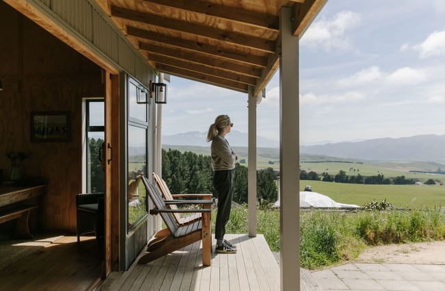 A lady standing on the wooden porch next to two desk chairs looking at the view.