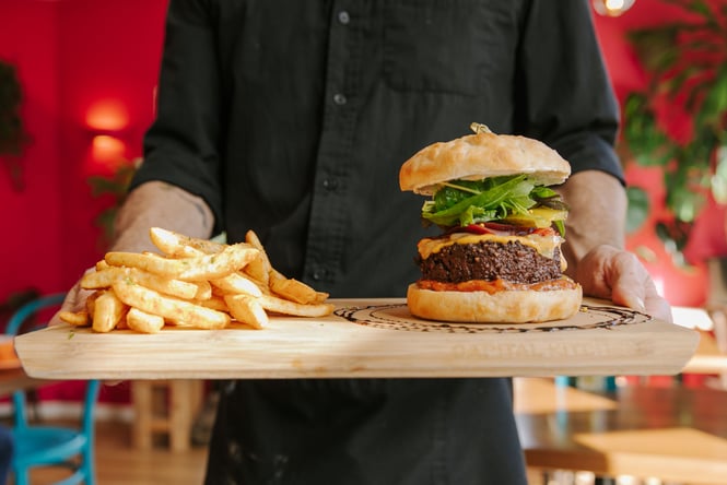 A person holding a wooden board featuring burger and a bowl of chips.